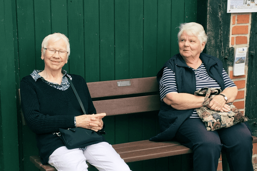 Zwei Frauen sitzen vor einer grüngestrichenen Holzwand auf einer Bank.