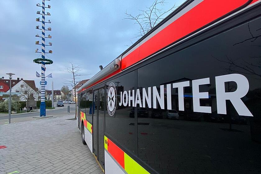 Ein langer Johanniter Bus steht auf einem gepflasterten Platz neben einem Maibaum.
