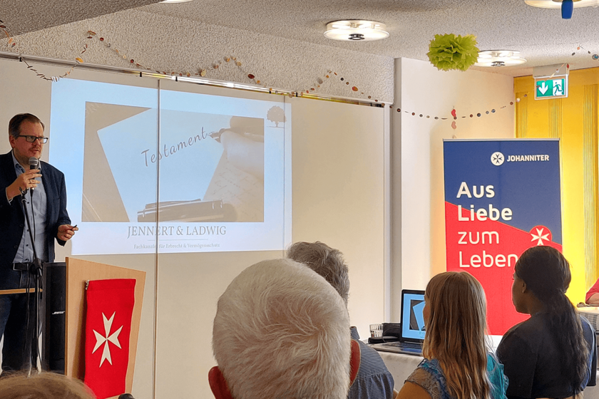 Links ein Mann im Anzug, er hält ein Mikrofon. Rechts einige Zuhörende von hinten zu sehen.