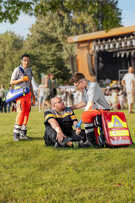 Ein, auf dem Boden einer Festwiese sitzender, Mann wird doch zwei Sanitäter versorgt. Im Hintergrund steht eine Menschenmenge vor einer großen Bühne.