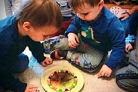 Mehrere Kinder blicken auf einen Teller mit Wasser und Süßigkeiten, die das Wasser bunt verfärben.