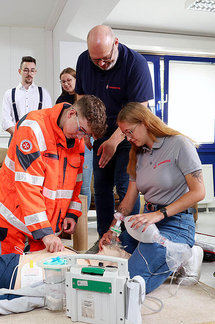 Eine Gruppe junger Menschen kniend an einer Übungspuppe. Ein Mann in Johanniter-Kleidung beugt sich zu ihnen. Andere schauen zu.