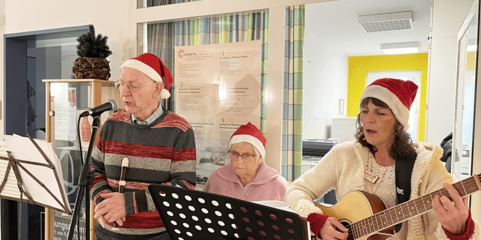 drei Personen mit Weihnachtsmützen stehen singen und mit Instrumenten hinter Notenständern